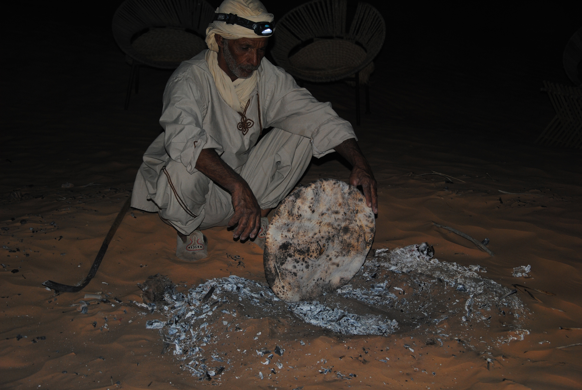 A Bedouin checks on bread baking under hot ashes in the Great Eastern Erg. Photo by me. Tuaregs and Bedouins traditionally bake a flatbread directly under hot ashes and sand in the desert. The Tuareg bread is called taguella (sometimes spelled tadjela or tagela), and is a Tuareg culinary staple. The dough, usually made from wheat, semolina, or millet, is shaped into a thick disc and buried in the embers, ashes, and sand of a small fire. It is cooked until done. Once cooked, the bread is cleaned of sand and ash and typically broken into pieces. It is then served with sauce or stew and often forms the centerpiece of a communal meal. This method is similar to the Bedouin technique for making arbood or abud bread. With this technique, a simple wheat dough is buried in the ashes and embers of a campfire and eaten with tea, ghee, or alongside meals. These traditions developed as adaptations to nomadic desert life, requiring no special oven or pan—just a fire and hot ashes.